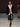 Look 04 desfile Un Romance Bien Latino 2026: saco sastre negro abierto con camisa blanca desabotonada sobre bermudas amplias de piel sintética negra, zapatos de vestir y detalle de rosetón en cintura.