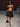 Look 06 desfile Un Romance Bien Latino 2026: top bandeau halter negro con tirantes desmontables y minifalda globo drapeada a tono, zapatillas de tacón con pulsera, bolso de rosetón textil y lentes de sol negros.