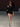 Look 12 desfile Un Romance Bien Latino 2026: vestido corto entallado negro de escote asimétrico y manga única acampanada con abertura, sandalias negras de tacón con pulsera, bolso de mano con rosetón textil y lentes de sol rectangulares.