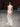 Look 13 desfile Un Romance Bien Latino 2026: vestido largo halter translúcido en gasa blanca con cuello alto fruncido y fajín a la cadera con rosetón textil al tono, aretes dorados colgantes.