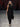 Look 18 desfile Un Romance Bien Latino 2026: vestido largo de fiesta en tejido de punto negro con escote asimétrico de hombro descubierto, cuello alto y manga capa envolvente, zapatillas negras de tacón.
