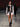 Look 19 desfile Un Romance Bien Latino 2026: chaleco motero de piel negra y camisa blanca abierta al pecho con rosetón negro y lazo colgante sobre shorts ajustados, botines chunky de piel negra.