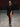 Look 21 desfile Un Romance Bien Latino 2026: enterizo asimétrico negro de mangas largas ceñido al cuerpo con una pierna descubierta y corte de tiro alto, sandalias negras de tacón decoradas con plumas y lentes de sol oscuros.