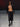 Look 23 desfile Un Romance Bien Latino 2026: bolero corto de piel negra con cuello chimenea y mangas largas abierto al pecho con bermudas sastre oversize negras de tiro medio, botines negros con tacón y plataforma y lentes de sol envolventes.